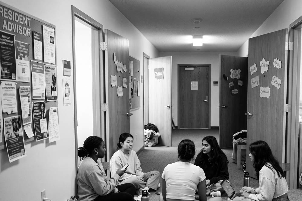 A dorm hallway in the evening, several doors propped open, a few students sitting on the floor of one room talking