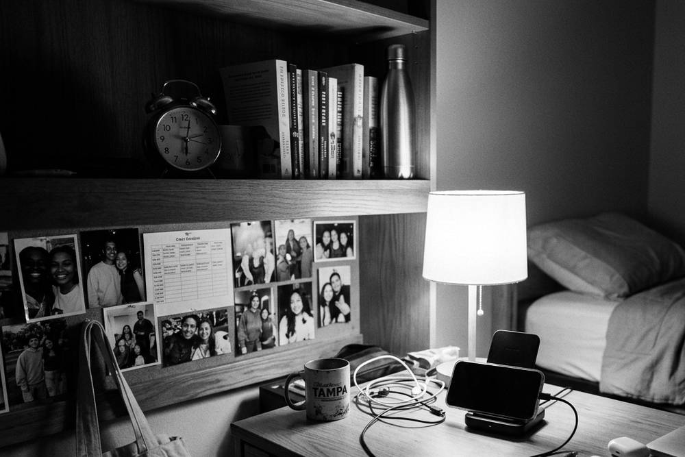 A dorm desk at night - phone parked in a charging station on the desk, alarm clock on the shelf, bed visible and screen-free in the background