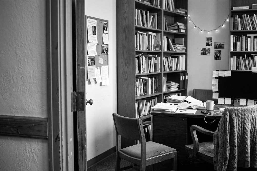 A professor's office in week two - door open, professor at a desk stacked with books, one empty chair waiting