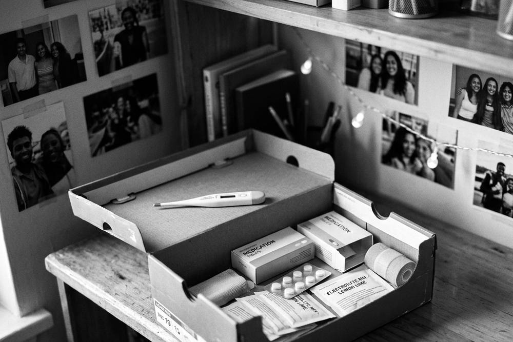 An open shoebox '2 a.m. kit' on a dorm shelf - thermometer, fever reducer, cold medicine, bandages, electrolyte packets