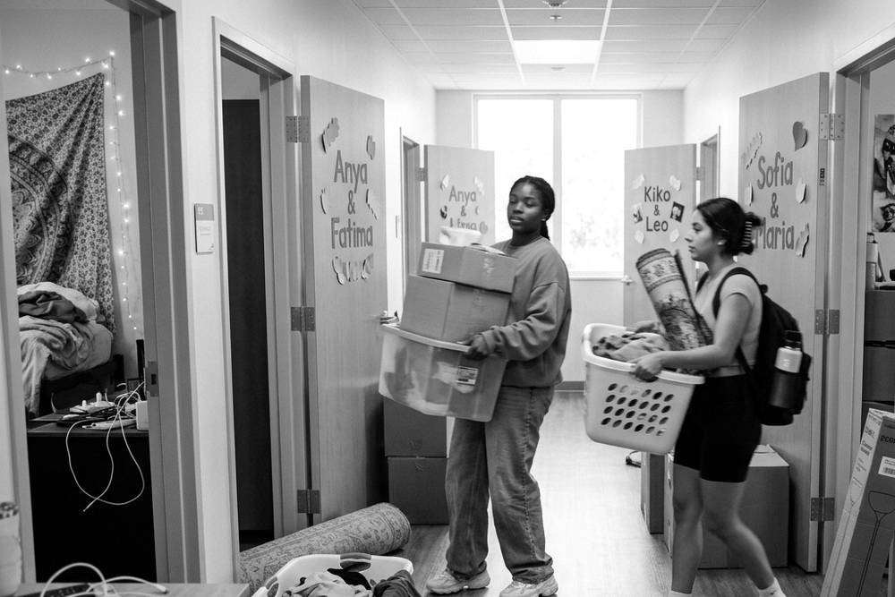 A dorm hallway in the first week, most doors propped open, light spilling across the floor, one door closed