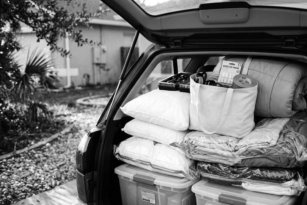 An open hatchback packed in labeled layers, a handwritten 'FIRST OUT' tote on top containing a tool kit, bedding, and command strips