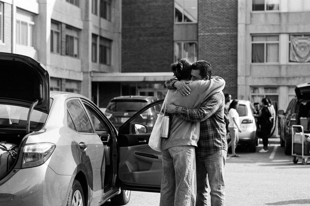 A parent and student in a quick hug by an open car door in a campus parking lot, dorm building in the background